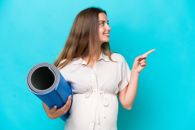 Jonge blanke vrouw geïsoleerd op blauwe achtergrond zwanger en houdt een mat vast terwijl ze iets presenteert