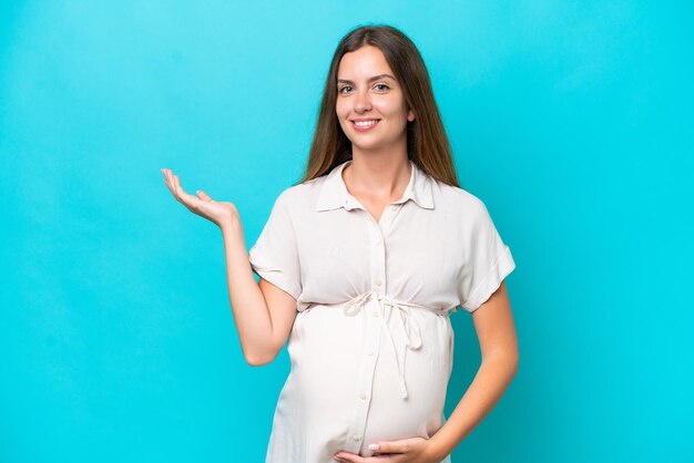 Jonge blanke vrouw geïsoleerd op blauwe achtergrond zwanger en iets presenteren