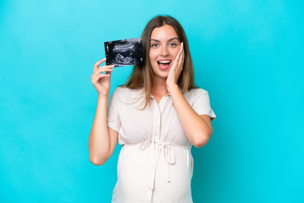 Jonge blanke vrouw geïsoleerd op blauwe achtergrond zwanger en verrast terwijl ze een echo vasthoudt