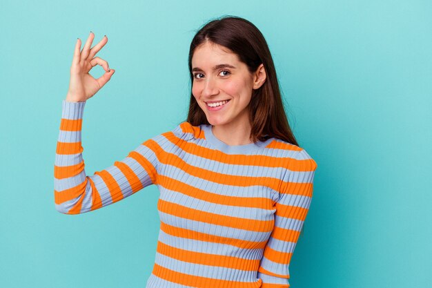 Jonge blanke vrouw geïsoleerd op blauwe muur knipoogt en houdt een goed gebaar met de hand.