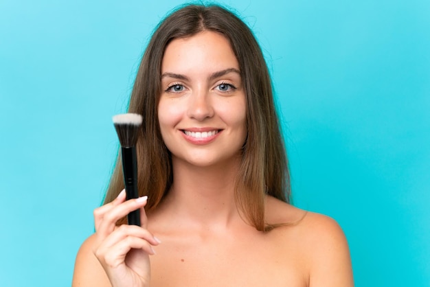 Jonge blanke vrouw geïsoleerd op een blauwe achtergrond met make-upborstel en een gelukkige uitdrukking