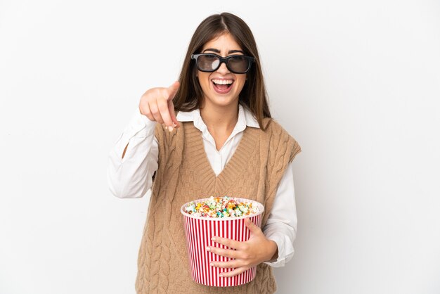 Jonge blanke vrouw geïsoleerd op een witte achtergrond met 3D-bril en houdt een grote emmer popcorn terwijl ze naar voren wijst