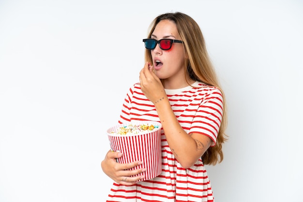 Jonge blanke vrouw geïsoleerd op een witte achtergrond met 3D-bril en met een grote emmer popcorns terwijl op zoek naar kant