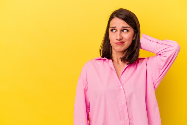 Jonge blanke vrouw geïsoleerd op gele achtergrond achterhoofd aanraken, denken en een keuze maken.