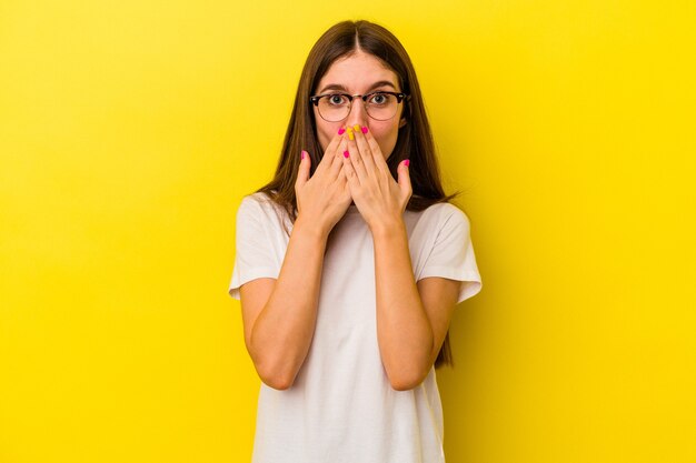 Jonge blanke vrouw geïsoleerd op gele achtergrond geschokt, mond bedekken met handen, angstig om iets nieuws te ontdekken.