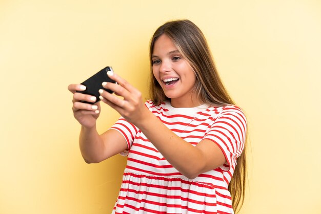 Jonge blanke vrouw geïsoleerd op gele achtergrond spelen met de mobiele telefoon