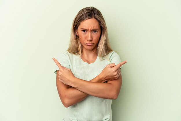 Jonge blanke vrouw geïsoleerd op groene achtergrond wijst zijwaarts, probeert te kiezen tussen twee opties.