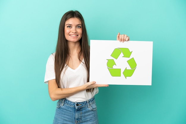 Jonge blanke vrouw geïsoleerd op het houden van een bordje met recycle icoon met gelukkige uitdrukking