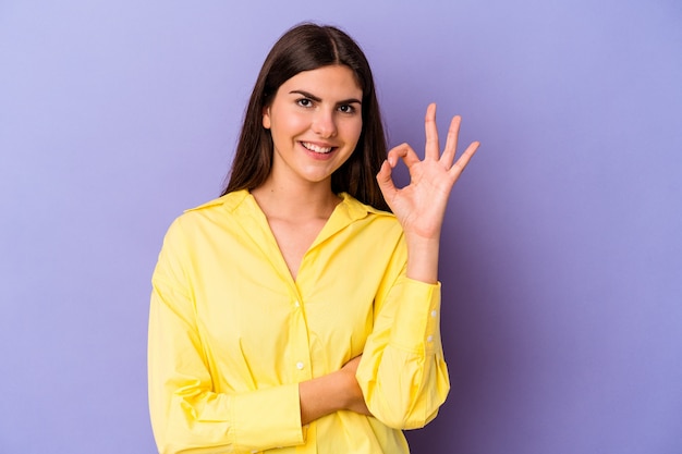 Jonge blanke vrouw geïsoleerd op paarse achtergrond knipoogt en houdt een goed gebaar met de hand.