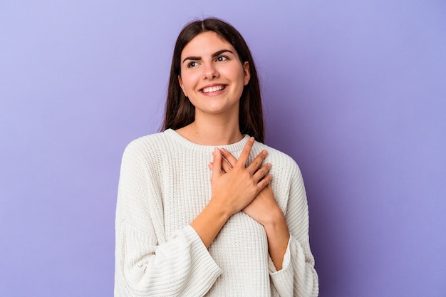 Jonge blanke vrouw geïsoleerd op paarse achtergrond lachen houden handen op hart, concept van geluk.