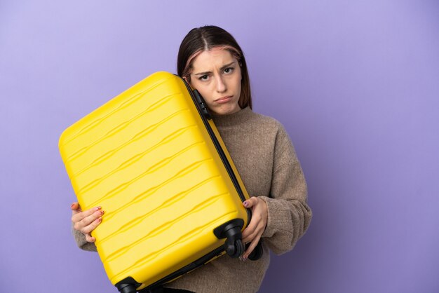 Jonge blanke vrouw geïsoleerd op paarse muur in vakantie met reiskoffer en ongelukkig