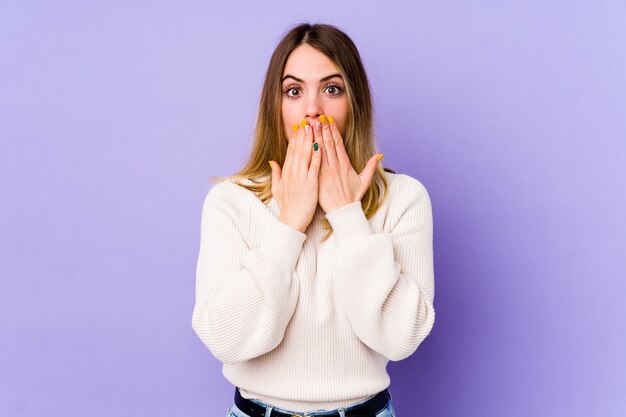 Jonge blanke vrouw geïsoleerd op paarse ruimte geschokt, mond bedekt met handen, angstig om iets nieuws te ontdekken.