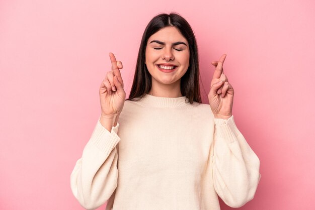 Jonge blanke vrouw geïsoleerd op roze achtergrond die vingers kruist om geluk te hebben