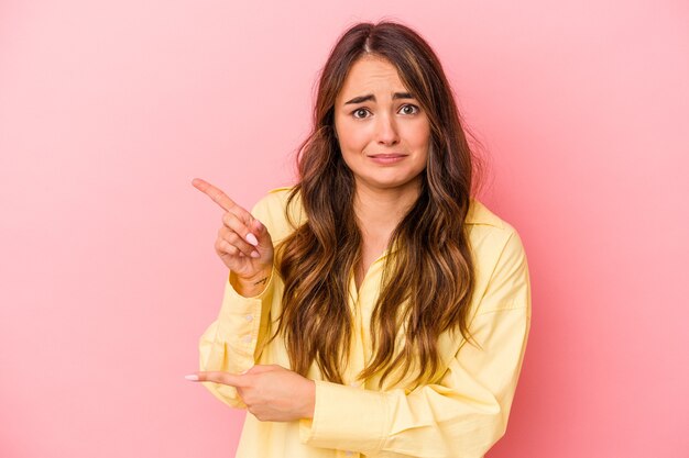 Jonge blanke vrouw geïsoleerd op roze achtergrond geschokt wijzend met wijsvingers naar een kopieerruimte.