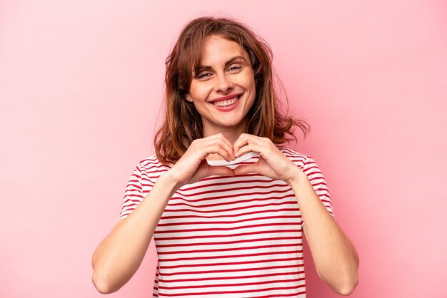 Jonge blanke vrouw geïsoleerd op roze achtergrond glimlachend en toont een hartvorm met handen