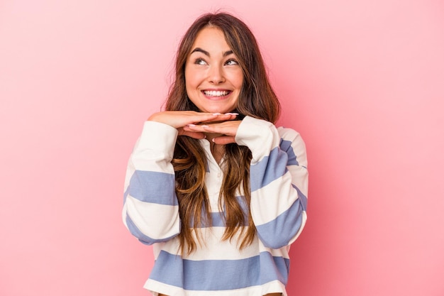 Jonge blanke vrouw geïsoleerd op roze achtergrond houdt handen onder de kin, kijkt gelukkig opzij.