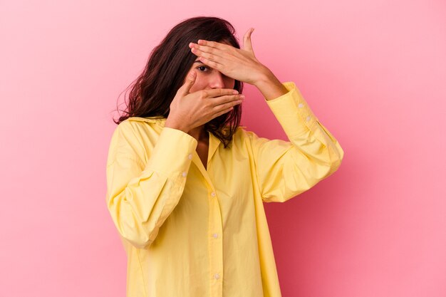 Jonge blanke vrouw geïsoleerd op roze achtergrond knippert naar de camera door vingers, beschaamd bedekkend gezicht.