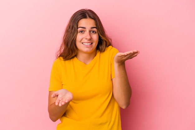 Jonge blanke vrouw geïsoleerd op roze achtergrond maakt schaal met armen, voelt zich gelukkig en zelfverzekerd.