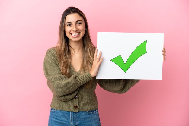 Jonge blanke vrouw geïsoleerd op roze achtergrond met een bordje met tekst Groen vinkje pictogram met gelukkige expressie