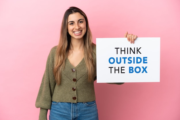 Jonge blanke vrouw geïsoleerd op roze achtergrond met een bordje met tekst Think Outside The Box met gelukkige uitdrukking