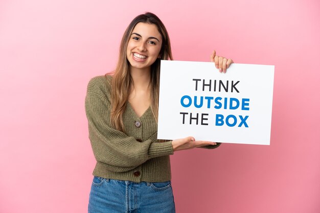 Jonge blanke vrouw geïsoleerd op roze achtergrond met een bordje met tekst Think Outside The Box met gelukkige uitdrukking