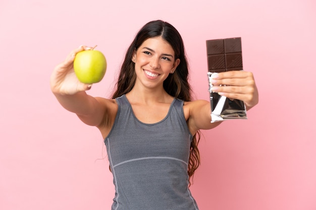 Jonge blanke vrouw geïsoleerd op roze achtergrond met een chocoladetablet in de ene hand en een appel in de andere