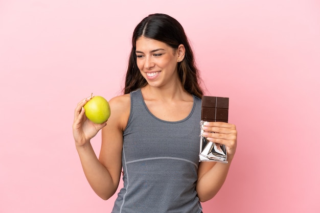 Jonge blanke vrouw geïsoleerd op roze achtergrond met een chocoladetablet in de ene hand en een appel in de andere