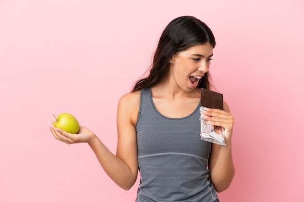 Jonge blanke vrouw geïsoleerd op roze achtergrond met een chocoladetablet in de ene hand en een appel in de andere