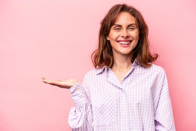 Jonge blanke vrouw geïsoleerd op roze achtergrond met een kopie ruimte op een handpalm en met een andere hand op de taille