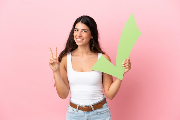 Jonge blanke vrouw geïsoleerd op roze achtergrond met een vinkje en een overwinning vieren