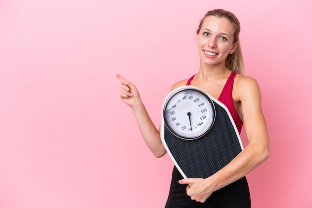 Jonge blanke vrouw geïsoleerd op roze achtergrond met weegmachine en wijzende kant