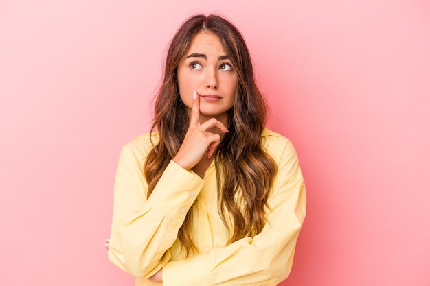 Jonge blanke vrouw geïsoleerd op roze achtergrond zijwaarts kijkend met twijfelachtige en sceptische uitdrukking.