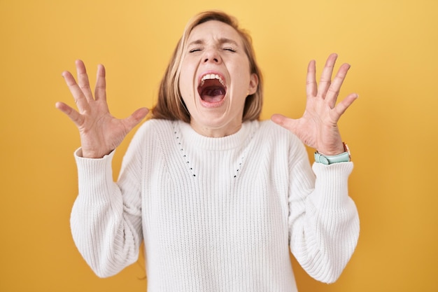 Jonge blanke vrouw, gekleed in een witte trui over een gele achtergrond, viert gek en gek voor succes met opgeheven armen en gesloten ogen opgewonden schreeuwend. winnaar concept