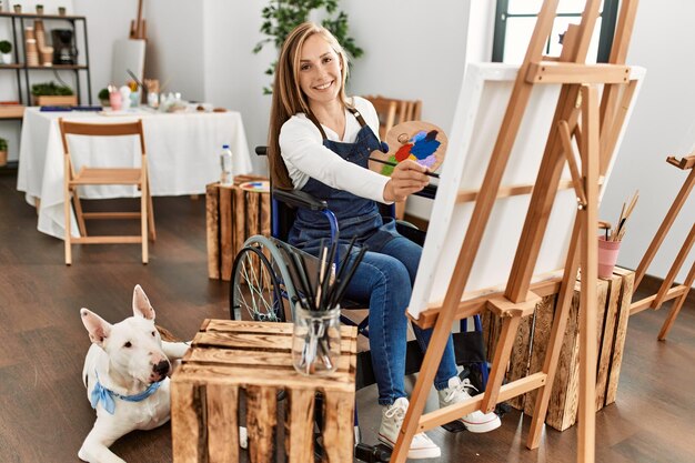 Jonge blanke vrouw glimlachend zelfverzekerd zittend op rolstoel tekenen met hond in kunststudio