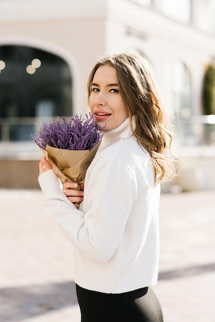 Jonge blanke vrouw houdt een paars boeket bloemen in haar handen en loopt langs een stadsstraat