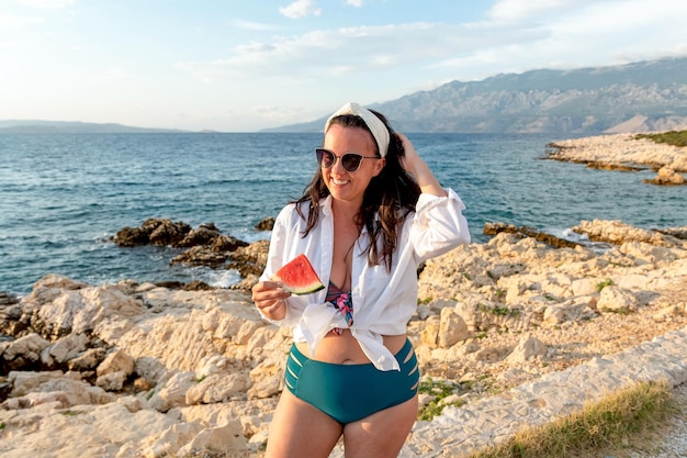 Jonge blanke vrouw in een badpak en zonnebril poseren met een stuk watermeloen op het strand