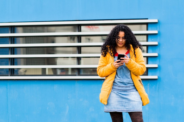 Jonge blanke vrouw in een denim overall jurk die naar haar telefoon kijkt op de achtergrond van een muur