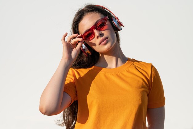Jonge blanke vrouw in een oranje t-shirt en rode zonnebril die naar muziek luistert met een koptelefoon