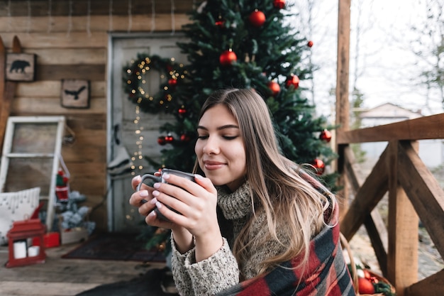 Jonge blanke vrouw in een plaid drinkt een warme drank op de veranda met kerstboom en decoraties.