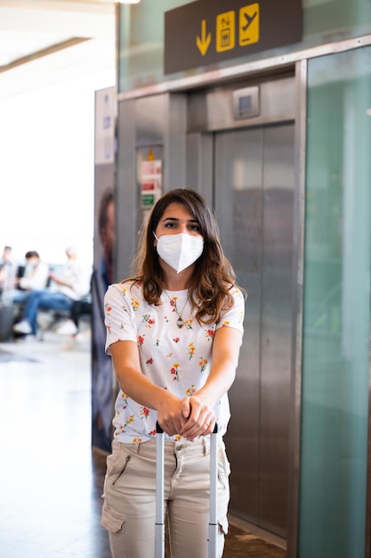 jonge blanke vrouw in vrijetijdskleding en gezichtsmasker die naar camera kijkt Vrouw met trolley