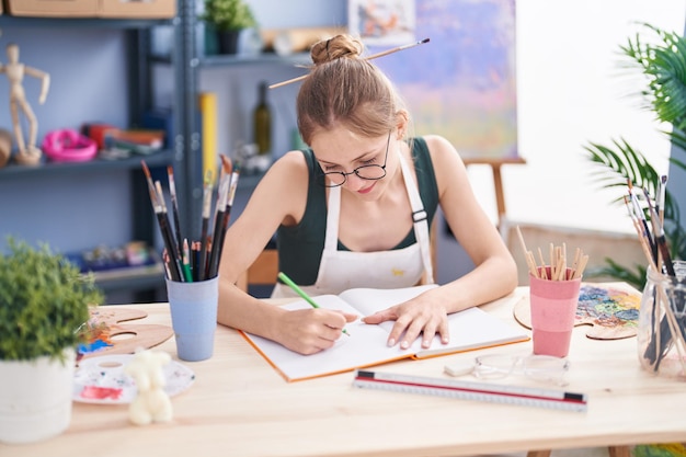 Jonge blanke vrouw kunstenaar glimlachend zelfverzekerd tekenen op notebook in kunststudio