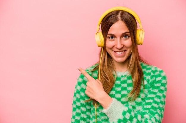 Jonge blanke vrouw luisteren naar muziek geïsoleerd op een roze achtergrond glimlachend en opzij wijzend met iets op lege ruimte