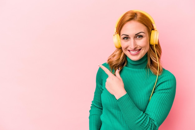 Jonge blanke vrouw luisteren naar muziek geïsoleerd op roze achtergrond glimlachend en opzij wijzend, iets laten zien op lege ruimte.