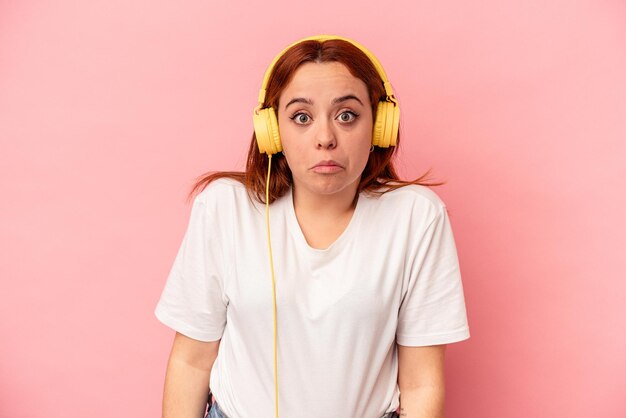 Jonge blanke vrouw luisteren naar muziek geïsoleerd op roze achtergrond haalt schouders op en open ogen verward.