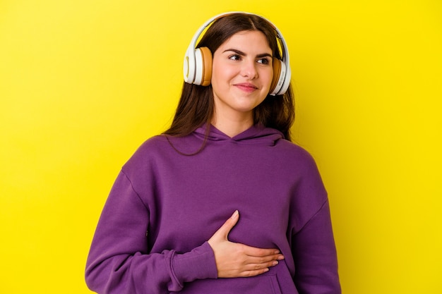 Jonge blanke vrouw luisteren naar muziek met een koptelefoon geïsoleerd op roze muur raakt buik, glimlacht zachtjes, eten en tevredenheid concept.