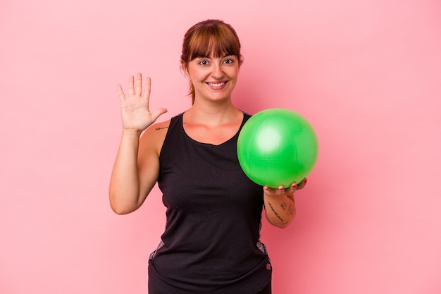 Jonge blanke vrouw met bal voor het doen van sport geïsoleerd op roze achtergrond glimlachend vrolijk met nummer vijf met vingers.