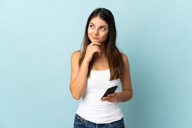 Jonge blanke vrouw met behulp van mobiele telefoon geïsoleerd op blauwe muur en opzoeken