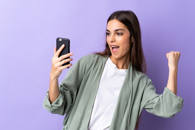 Jonge blanke vrouw met behulp van mobiele telefoon geïsoleerd op paarse achtergrond een overwinning vieren