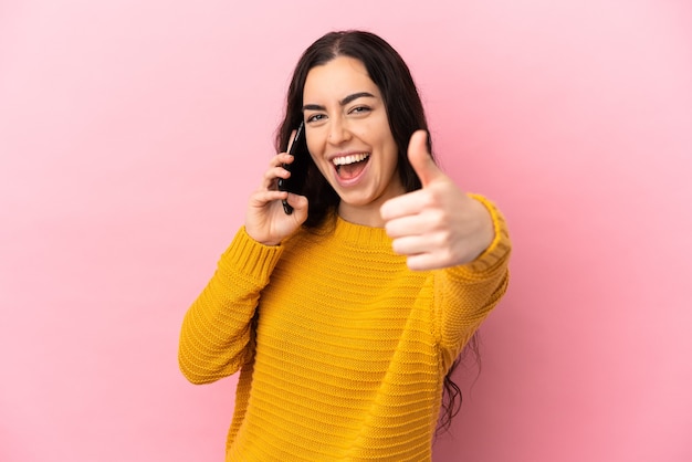 Jonge blanke vrouw met behulp van mobiele telefoon geïsoleerd op roze achtergrond met duimen omhoog omdat er iets goeds is gebeurd