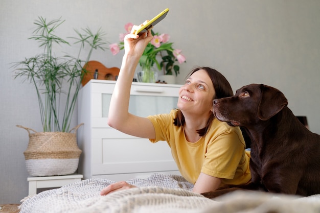 Jonge blanke vrouw met donker haar in een gele t-shirt ligt op een bed met een hond